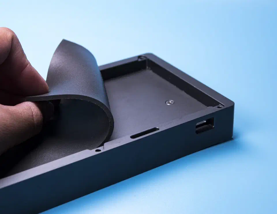 A hand lowering a black Poron foam sheet into the bottom of an empty dark grey aluminium keyboard case
