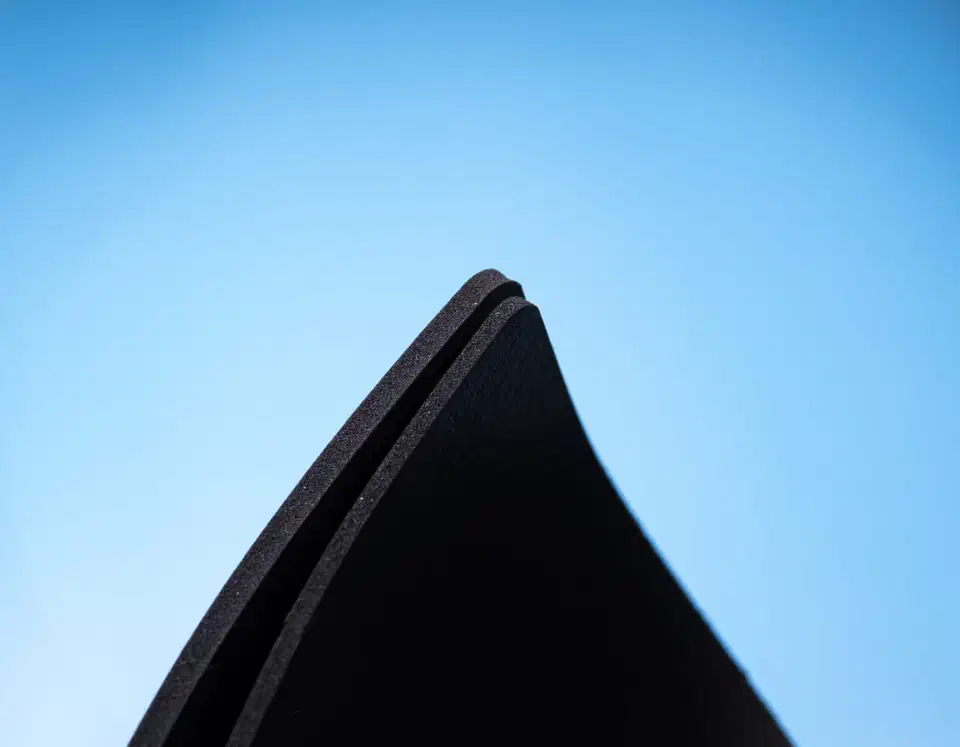 Close-up of the edges of two stacked black Poron foam sheets curving upward against a blue background, showing their fine open-cell texture