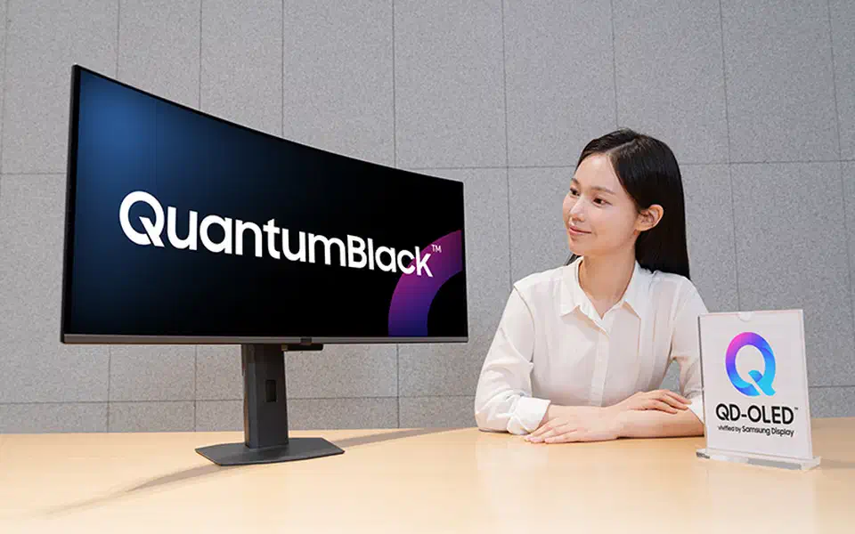 Ultrawide QD-OLED monitor in a bright studio showing QuantumBlack on a deep black screen, with a QD-OLED verified sign on the desk